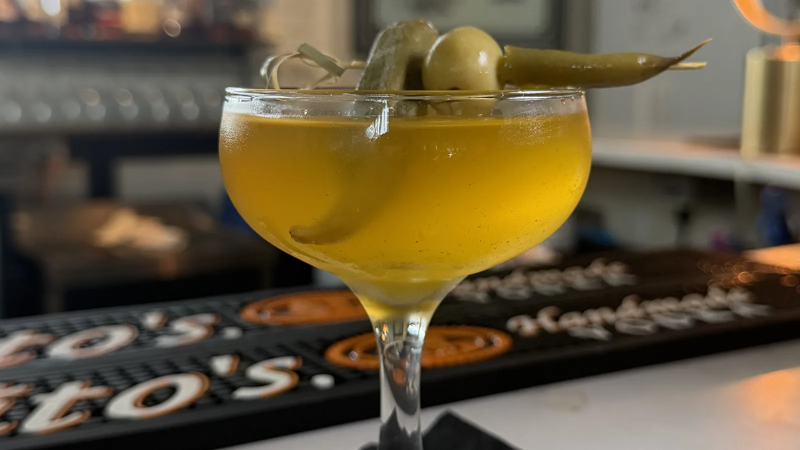 The Cajun Dirty Martini at Roch's Reprieve inside St. Roch Market, a golden cocktail in a coupe garnished with pickled okra and olives, sitting on a bar mat