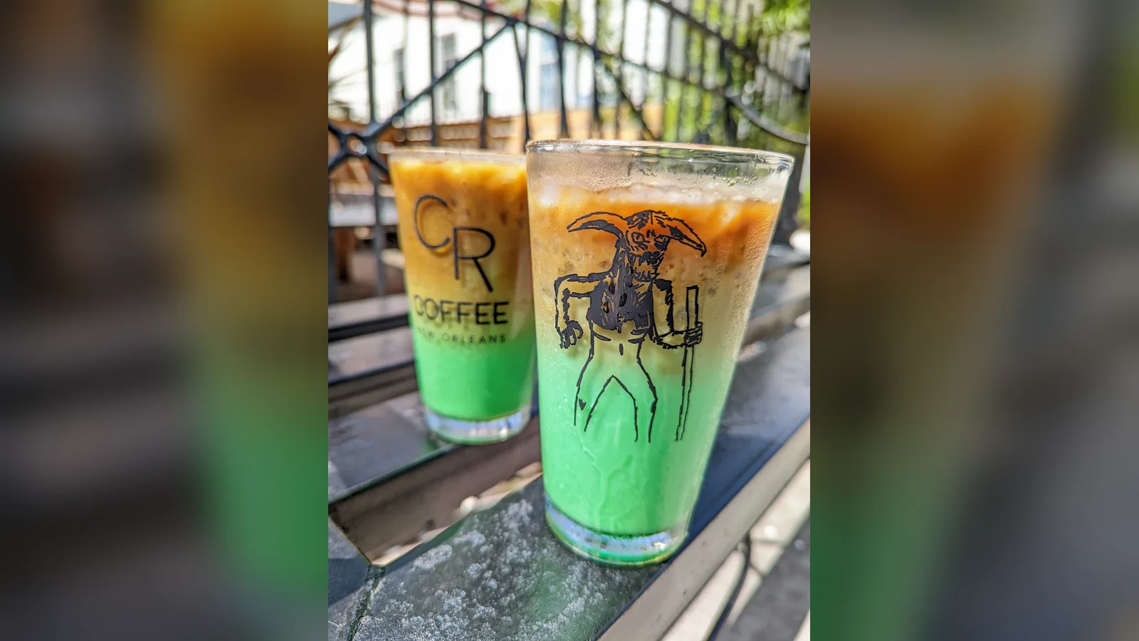Two iced pandan lattes from CR Coffee on a metal rail, bright green at the bottom under a layer of coffee, one glass printed with the Bayou Beast illustration and one with the CR Coffee logo