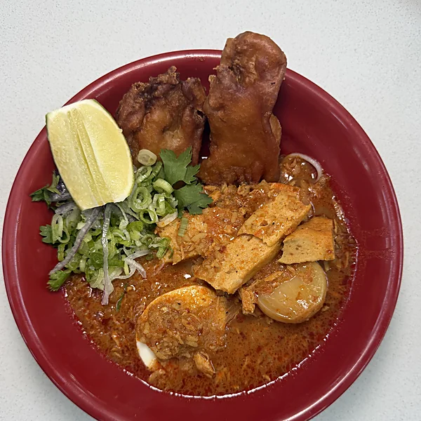 A red bowl of mohinga at Laksa with fried fritters, a lime wedge, scallions, tofu, and a halved boiled egg in deep red broth