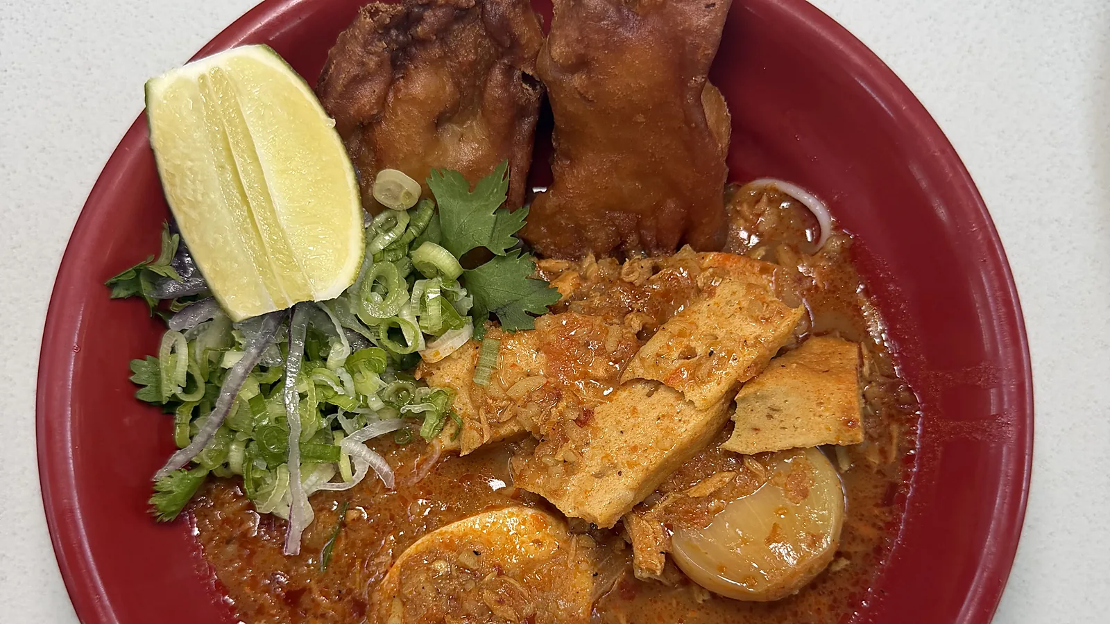 A red bowl of mohinga at Laksa: rice noodles in a deep red broth with fried fritters, a lime wedge, sliced scallions, cilantro, slabs of tofu, and a halved boiled egg