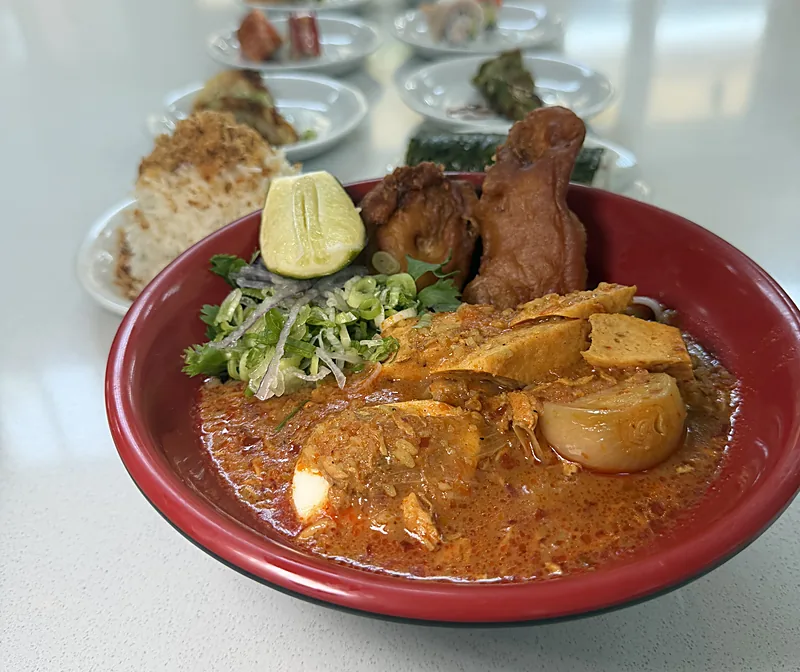 The mohinga bowl at Laksa on a marble counter inside St. Roch Market, with plates of sushi, rice, and greens out of focus behind it
