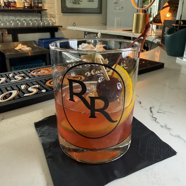 A rum Old Fashioned in a Roch's Reprieve RR-logo glass with a brandied cherry and orange peel over ice