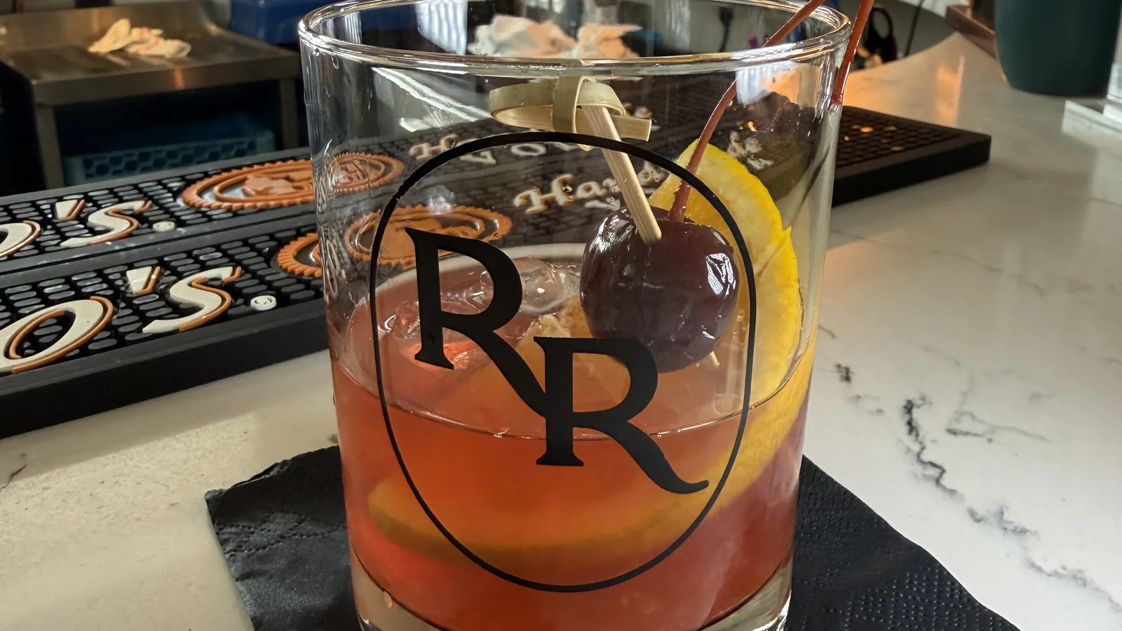 A rum Old Fashioned in a Roch's Reprieve rocks glass marked with the RR logo, garnished with a brandied cherry and an orange peel over ice, on a black napkin on a marble bar